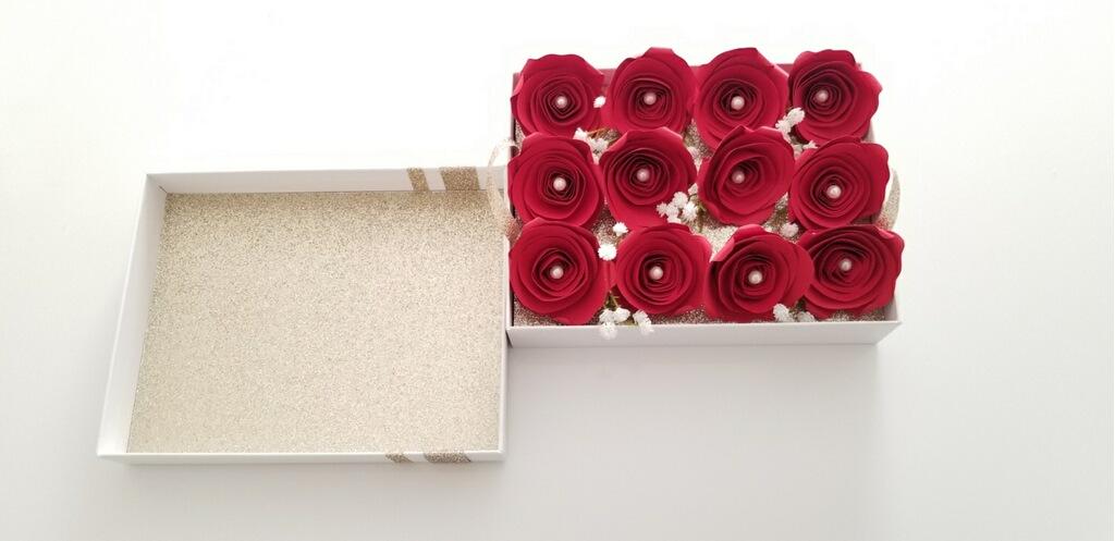 A full view of the one dozen red, paper roses sitting atop the removable tray inside the box; pictured with the inside of the lid.