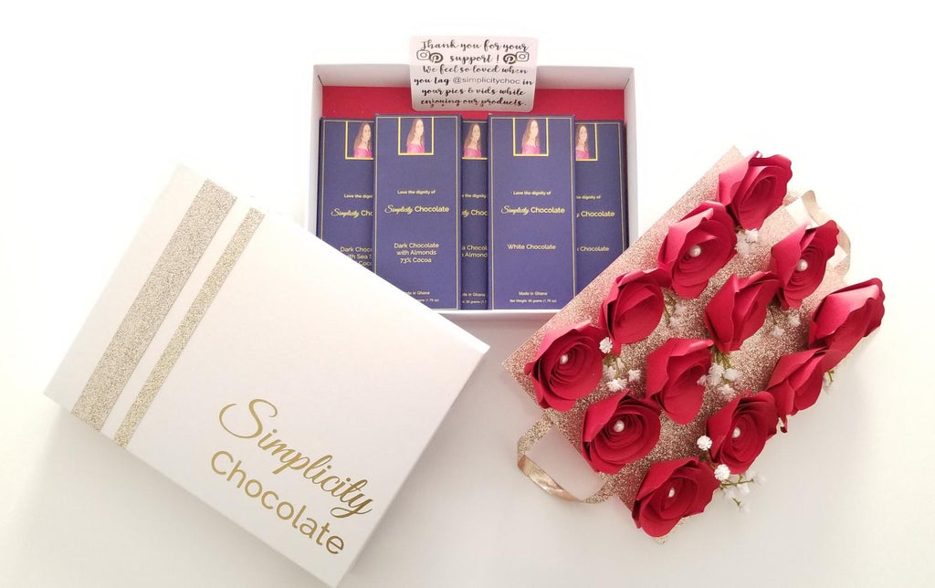 The Simplicity Chocolate Nostalgia Box is seen here as a white box with gold accents. The Simplicity Chocolate signature logo adorns the lid in gold print. A layer of one dozen red, paper roses sits atop a gold platform with handles that when lifted reveal the five gourmet Simplicity Chocolate flavor offerings nestled in the keepsake storage portion of the box.