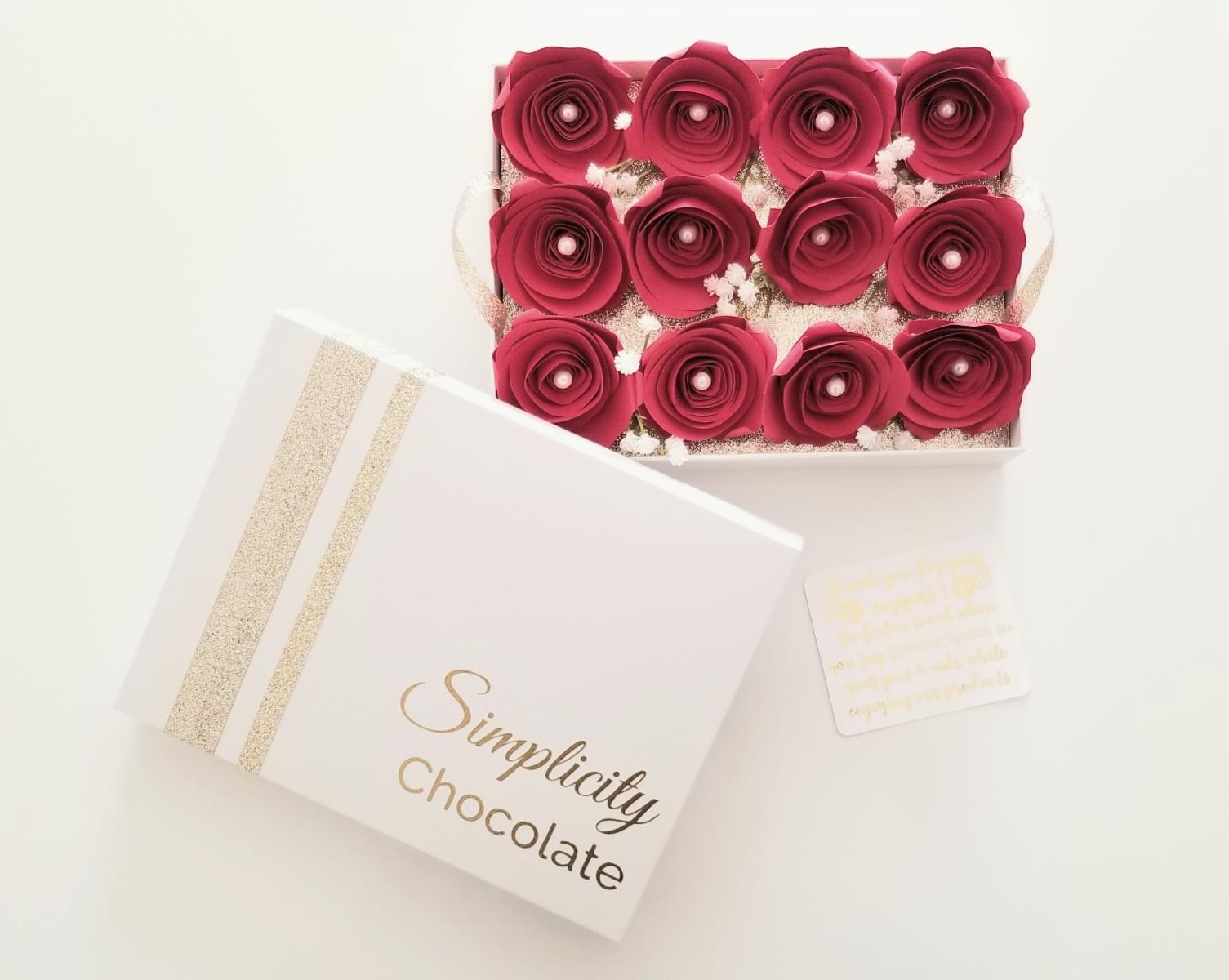 The lid of The Simplicity Chocolate Nostalgia Box with gold accents and the signature logo in gold writing is pictured next to a full view of the one dozen red, paper roses sitting atop the removable tray inside the box.