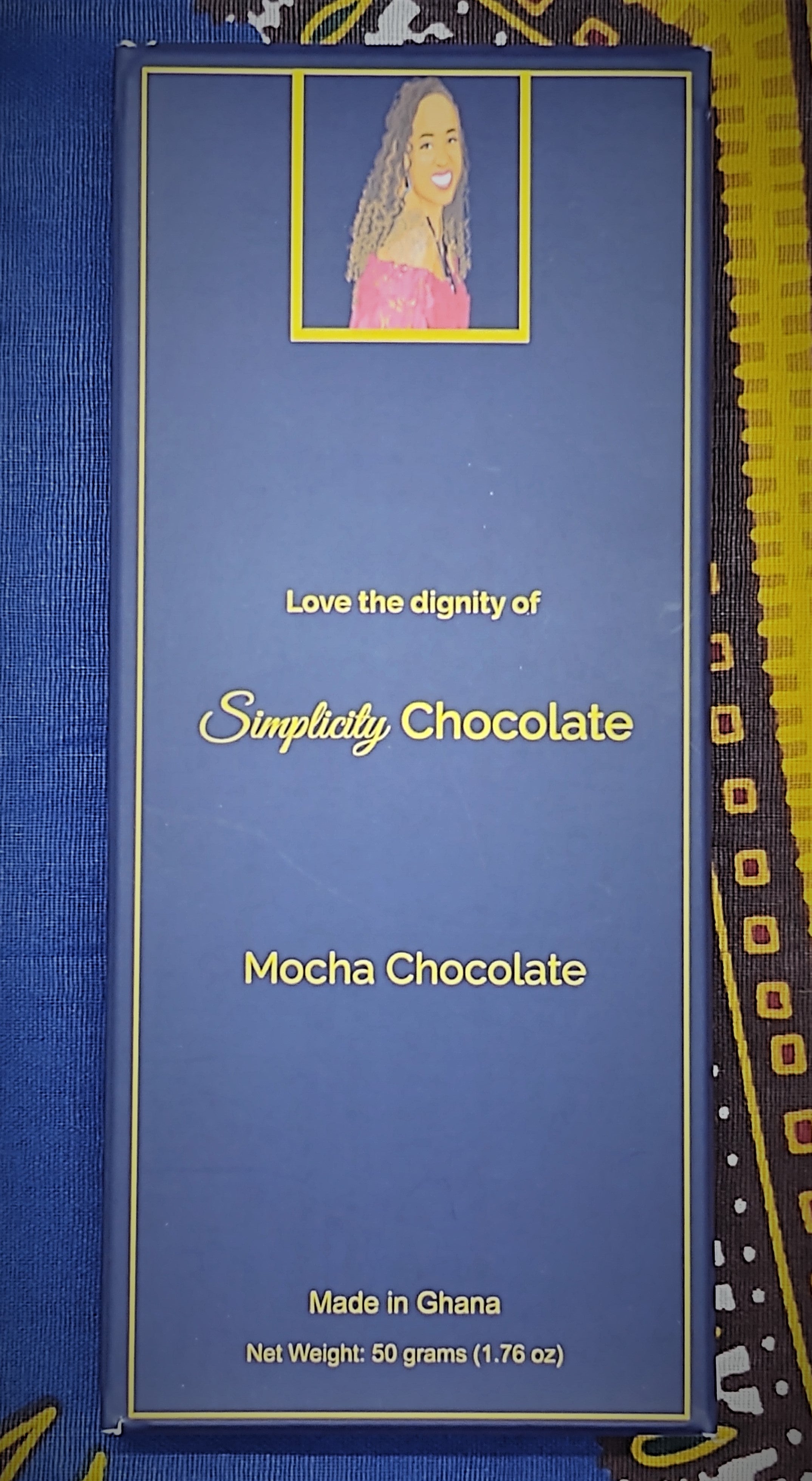Pictured: The Simplicity Chocolate bar wrapped in complete packaging which consists of a dark blue box with gold writing. A portrait of the CEO is centered at the top of the packaging. The CEO is seated, looking over her right shoulder, wearing a magenta, off the shoulder top and a royal blue gemstone necklace with matching earrings. The packaging reads, "Love the dignity of Simplicity Chocolate" in the center of the package. Mocha Chocolate. Made in Ghana. Net Weight: 50 grams (1.76 oz).