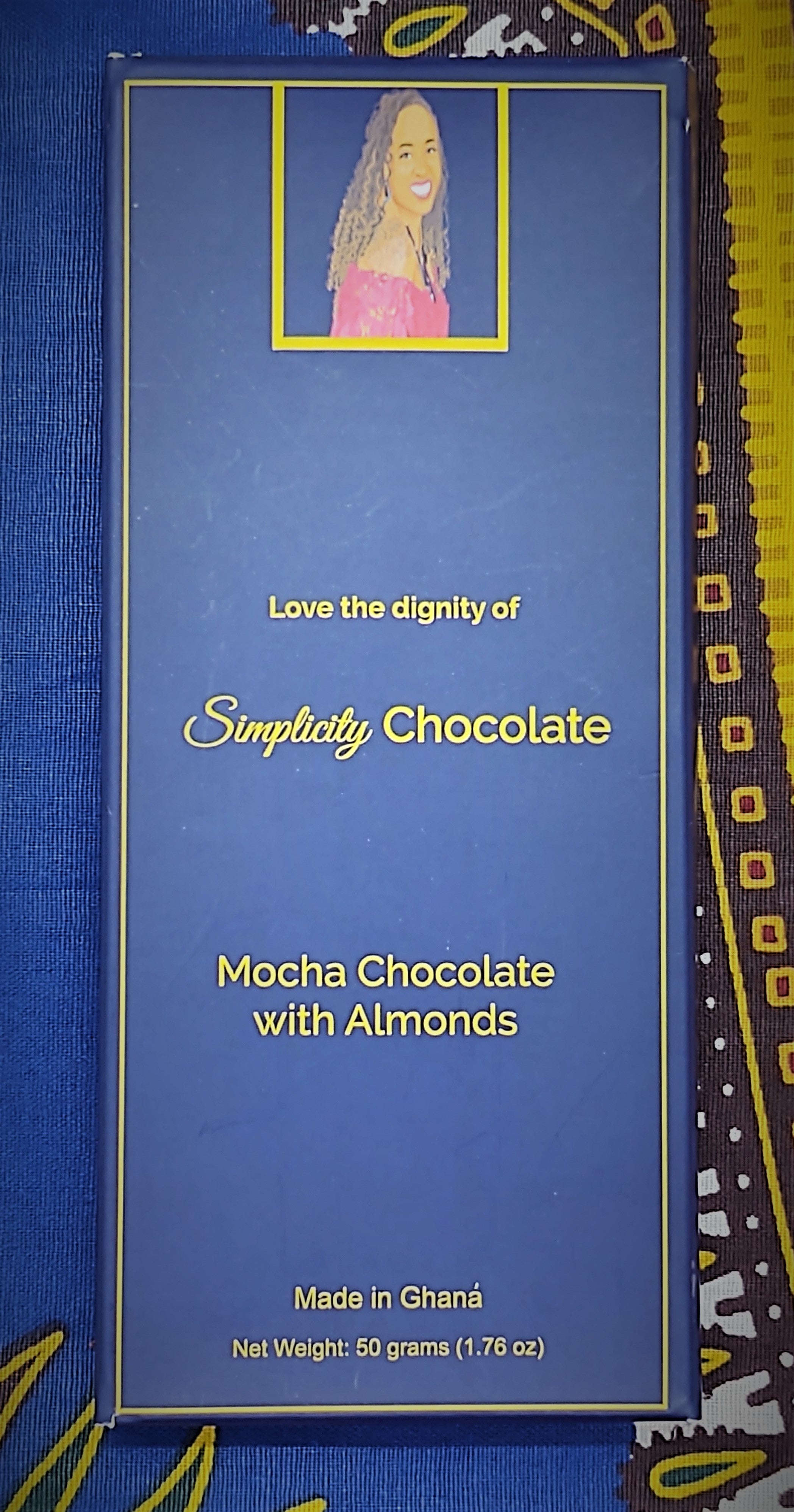 Pictured: The Simplicity Chocolate bar wrapped in complete packaging which consists of a dark blue box with gold writing. A portrait of the CEO is centered at the top of the packaging. The CEO is seated, looking over her right shoulder, wearing a magenta, off the shoulder top and a royal blue gemstone necklace with matching earrings. The packaging reads, "Love the dignity of Simplicity Chocolate" in the center of the package. Mocha Chocolate with Almonds. Made in Ghana. Net Weight: 50 grams (1.76 oz).