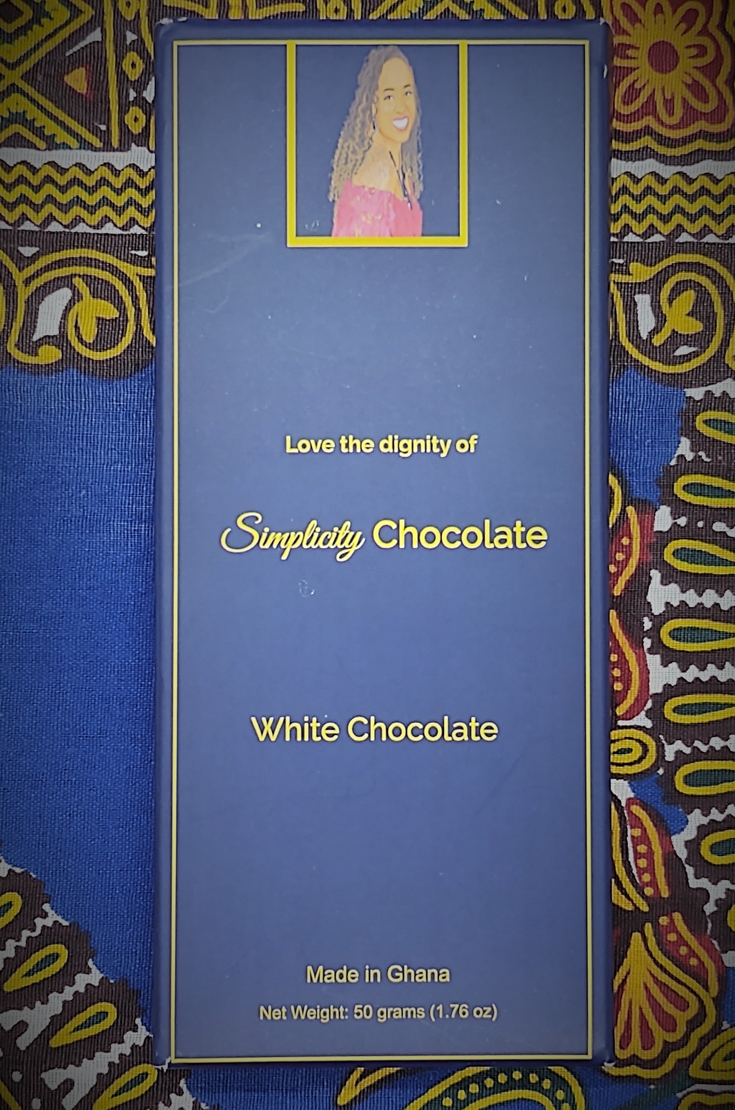 Pictured: The Simplicity Chocolate bar wrapped in complete packaging which consists of a dark blue box with gold writing. A portrait of the CEO is centered at the top of the packaging. The CEO is seated, looking over her right shoulder, wearing a magenta, off the shoulder top and a royal blue gemstone necklace with matching earrings. The packaging reads, "Love the dignity of Simplicity Chocolate" in the center of the package. White Chocolate. Made in Ghana. Net Weight: 50 grams (1.76 oz).
